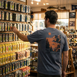 Point-Delgada-6.1oz-Heavyweight-Graphic-Tee-Steely-Grey-Back-View-Tackle-Shop. A maritime professional viewed from the back, wearing the steely grey Point Delgada 100% ring-spun cotton graphic tee featuring the archival 'Lost Coast' F/V Ironwright design. The subject is inspecting a pegboard display of commercial fishing lures in an authentic, well-stocked tackle store under warm natural light, highlighting the garment's relaxed fit, crisp graphic print, and durable double-needle topstitched construction.